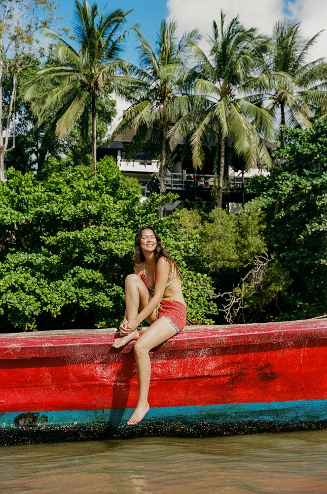 Woman sitting on a red boat in a tropical setting with palm trees and greenery.