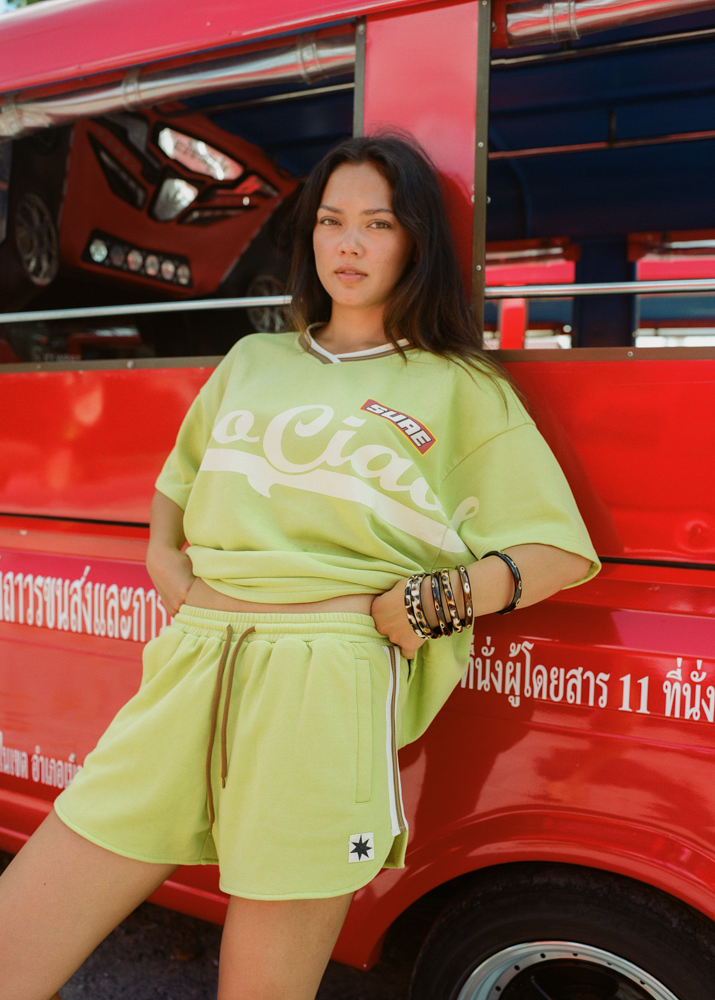 Woman in a bright green lounge jersey and drawstring shorts with CCV branding and logos standing in front of a red vehicle