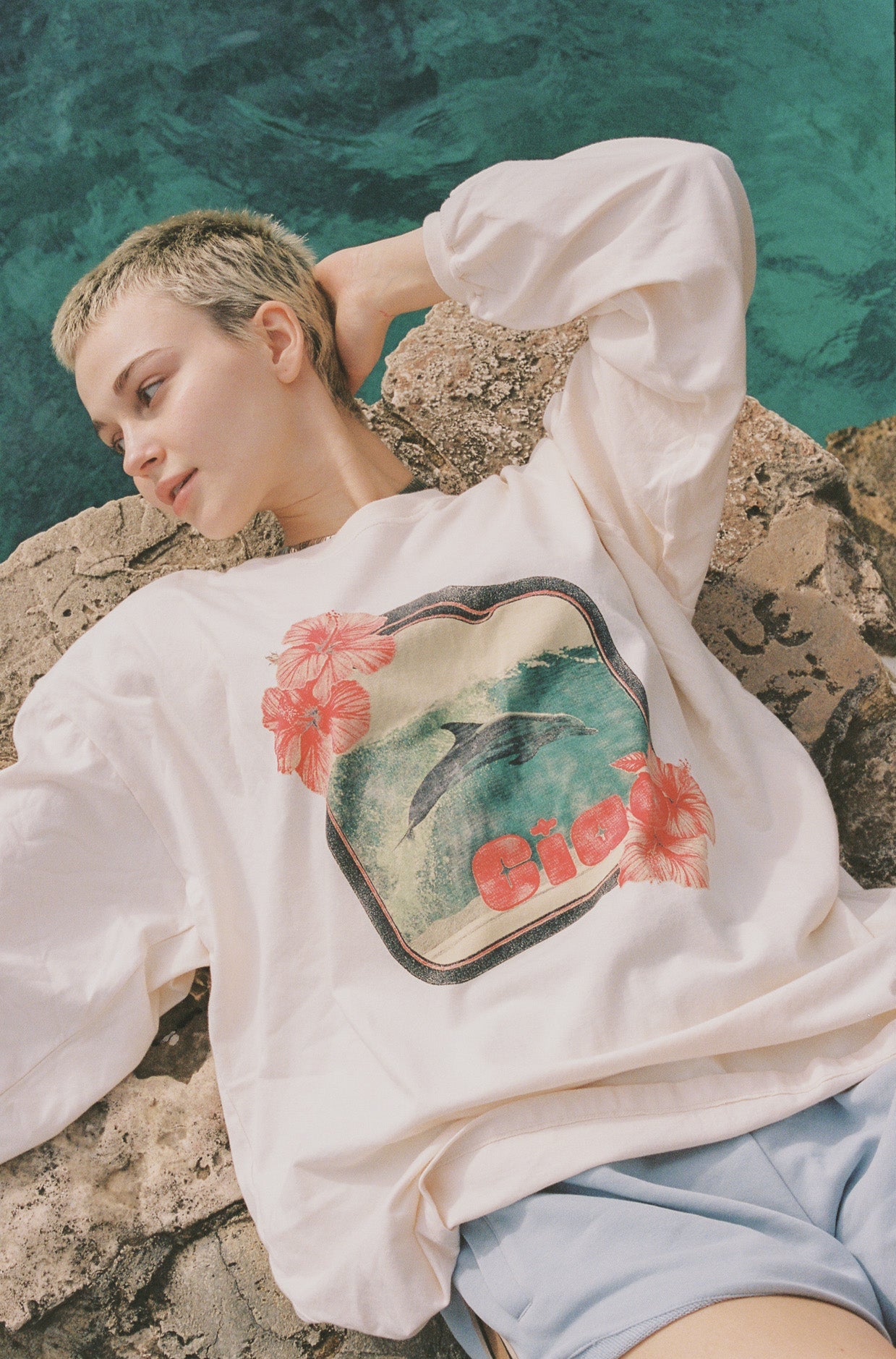 Person wearing a white long sleeve t-shirt with a colorful graphic dolphin design, standing on a rocky surface with a teal background.
