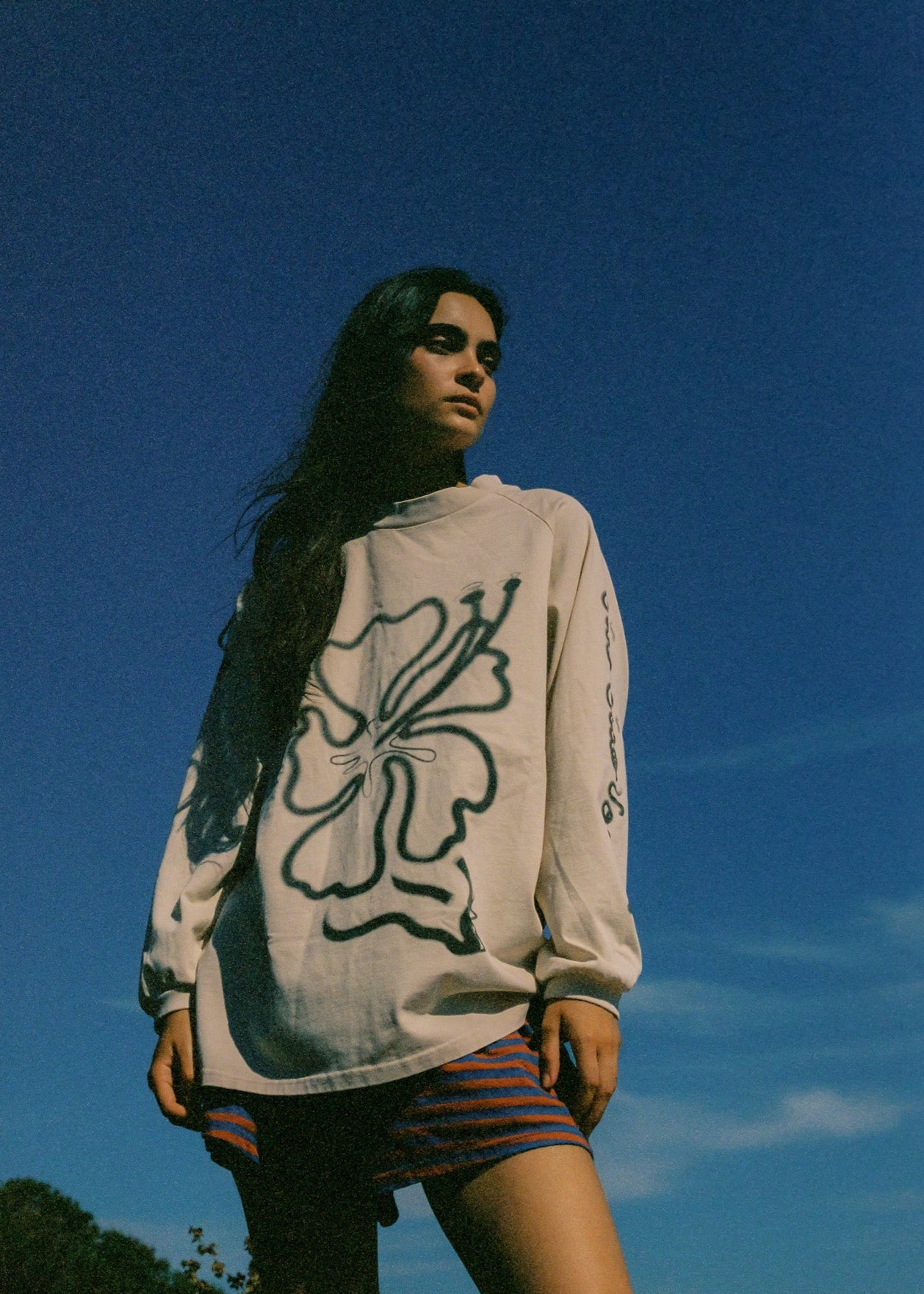 Person wearing a long sleeve t-shirt with a hibiscus logo and striped shorts along a blue sky background.