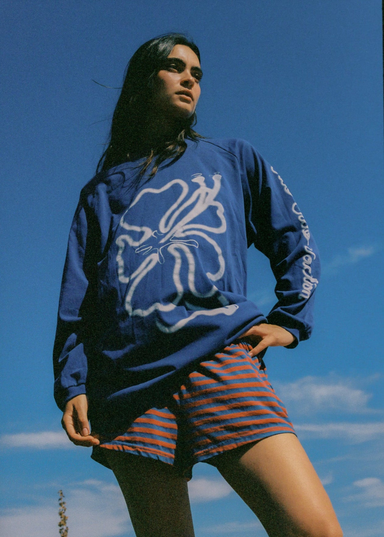 Person wearing a blue oversized long sleeve t-shirt with a white hibiscus design and striped shorts against a blue sky.