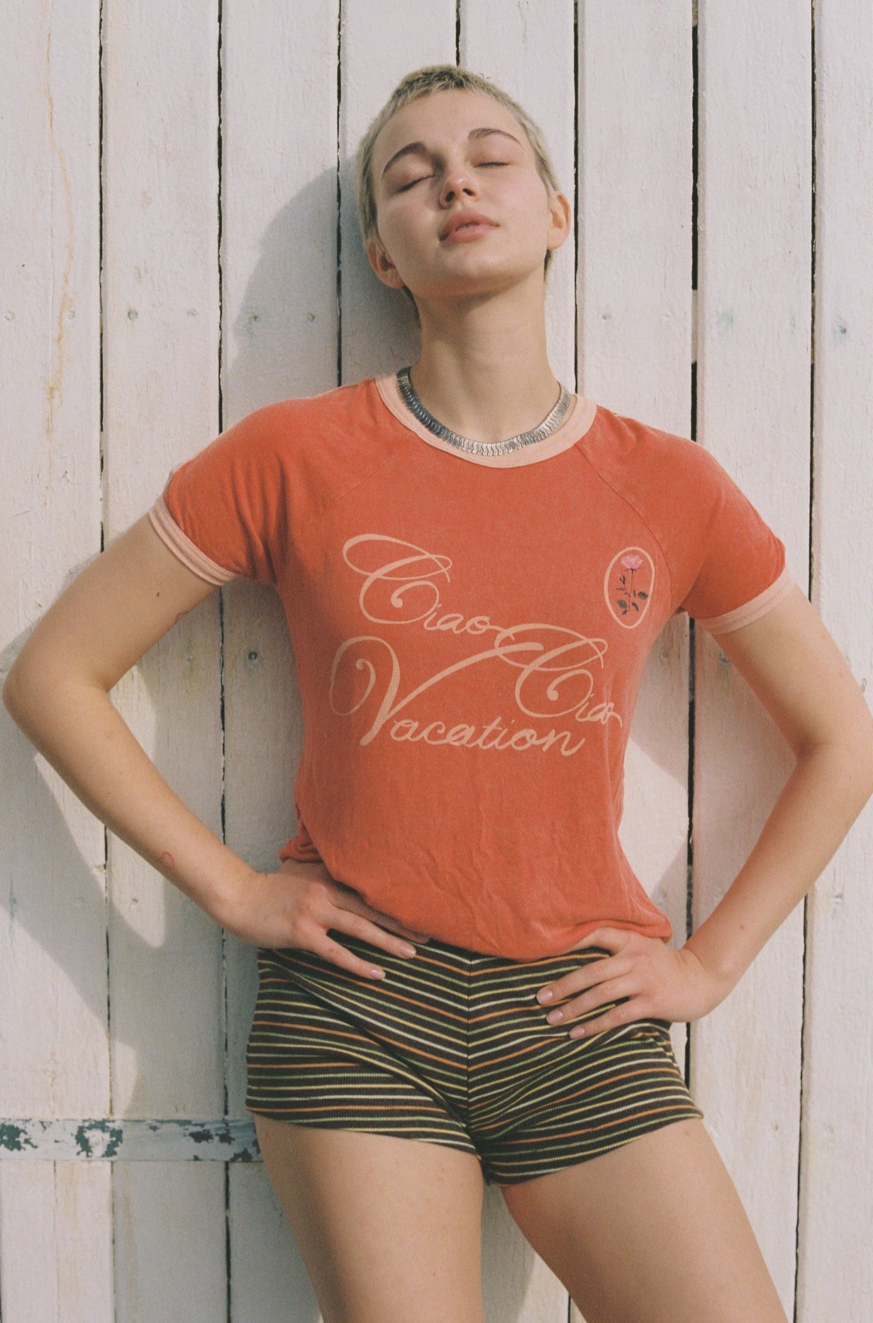 Person wearing an red t-shirt with pink Ciao Ciao Vacation text and striped shorts against a wooden background