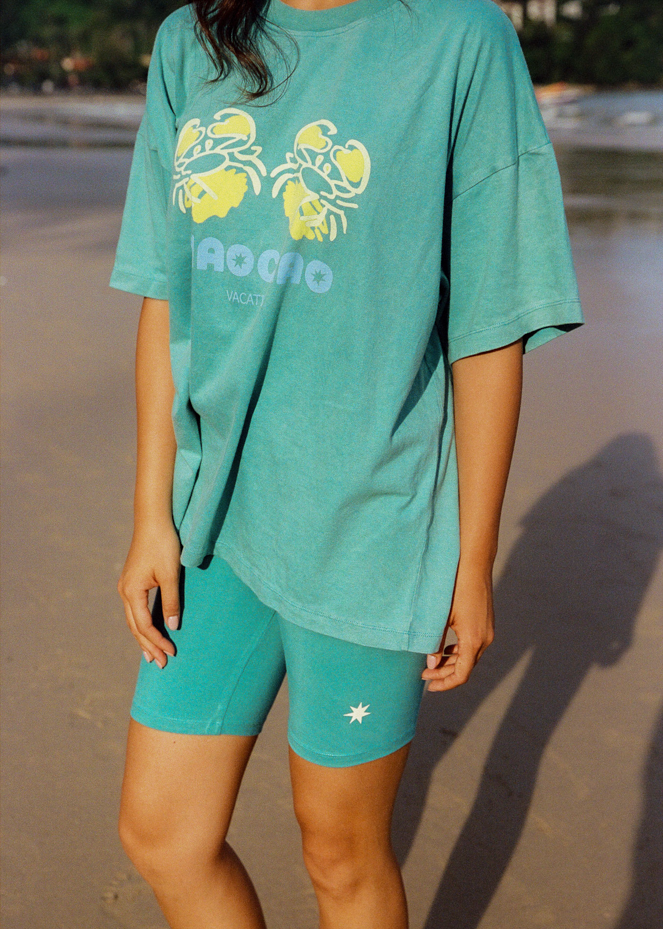 Woman in teal oversized t-shirt with a green crab graphic design and "Ciao Ciao" branding on a beach 