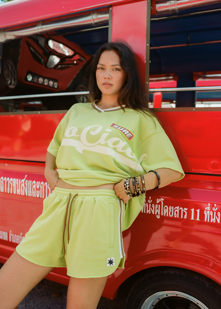 Woman wearing a bright green oversized jersey t-shirt with 'Ciao Ciao' text, sitting outside a vehicle.