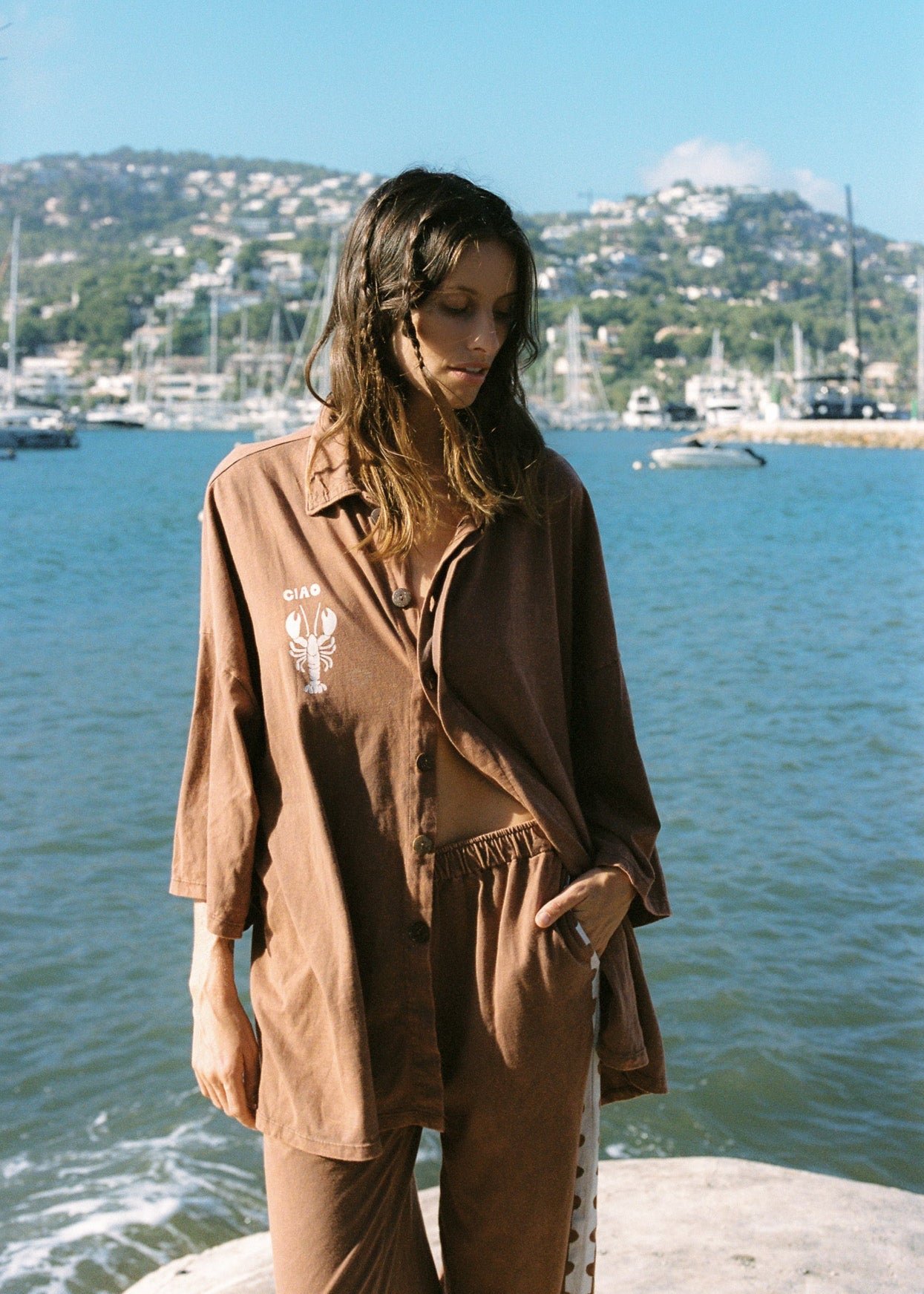 A person standing by the water wearing a brown oversized bungalow button up shirt.