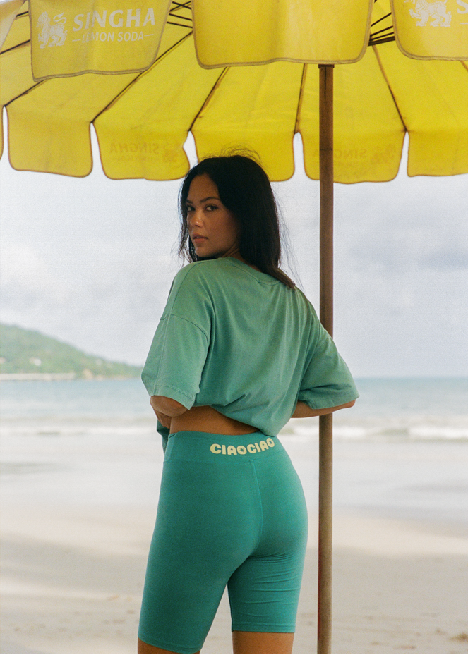 Woman in blue bike short with "CIAO" logo and blue oversized t-shirt on a beach standing under a yellow umbrella on a beach