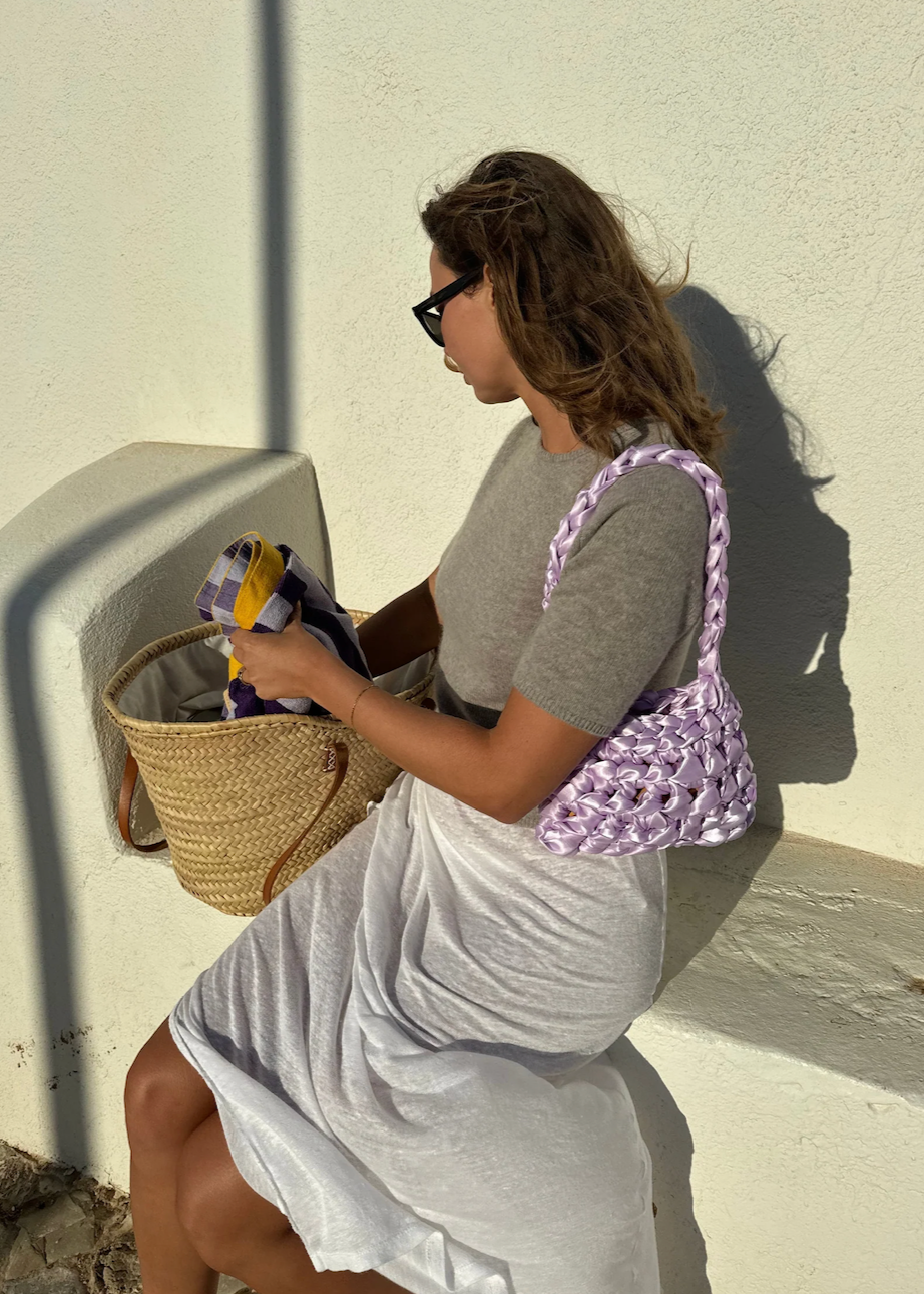 Woman sitting against a white wall wearing a purple satin knot woven shoulder bag
