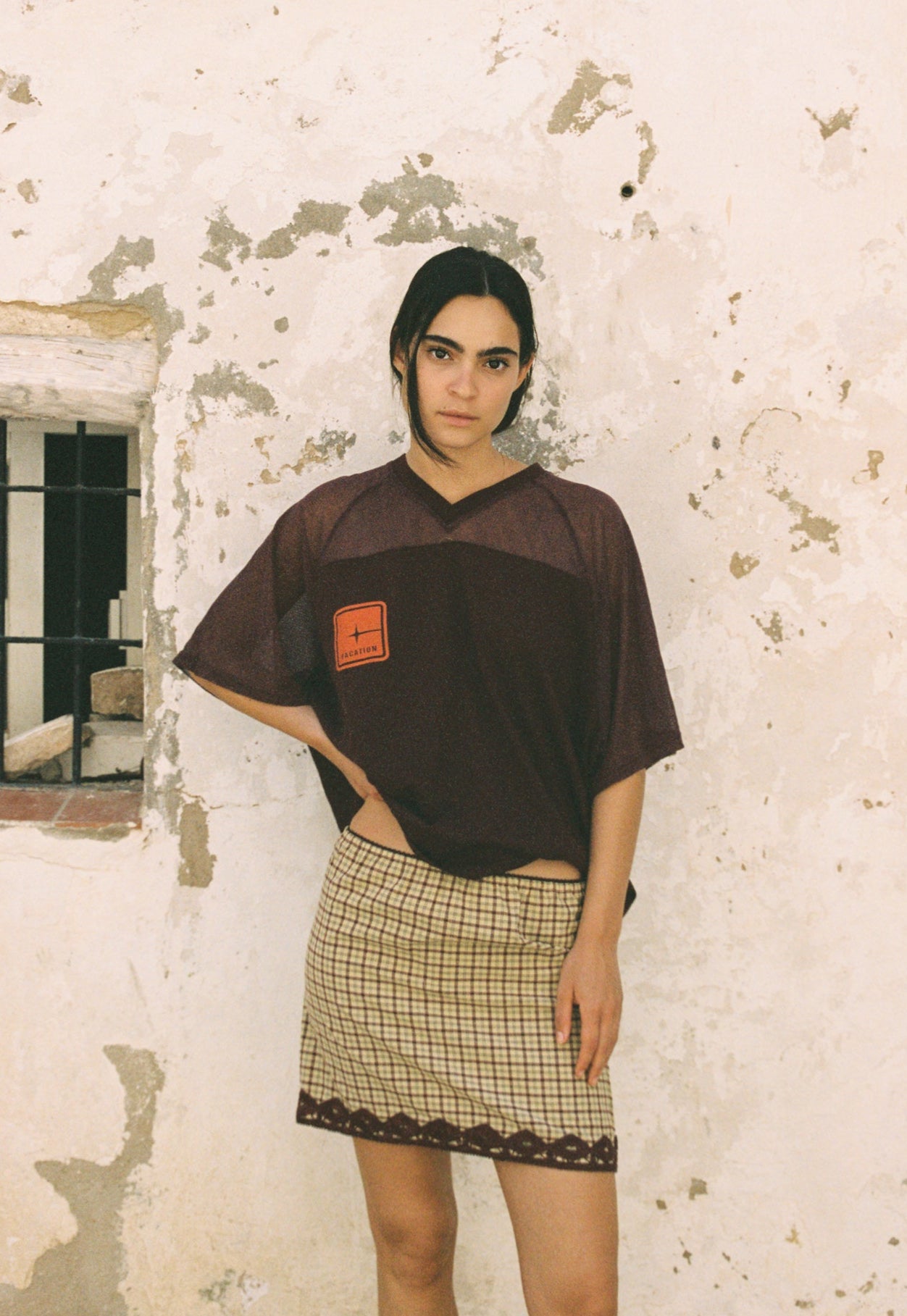 Woman wearing a brown oversized t-shirt and plaid skirt against a textured wall.