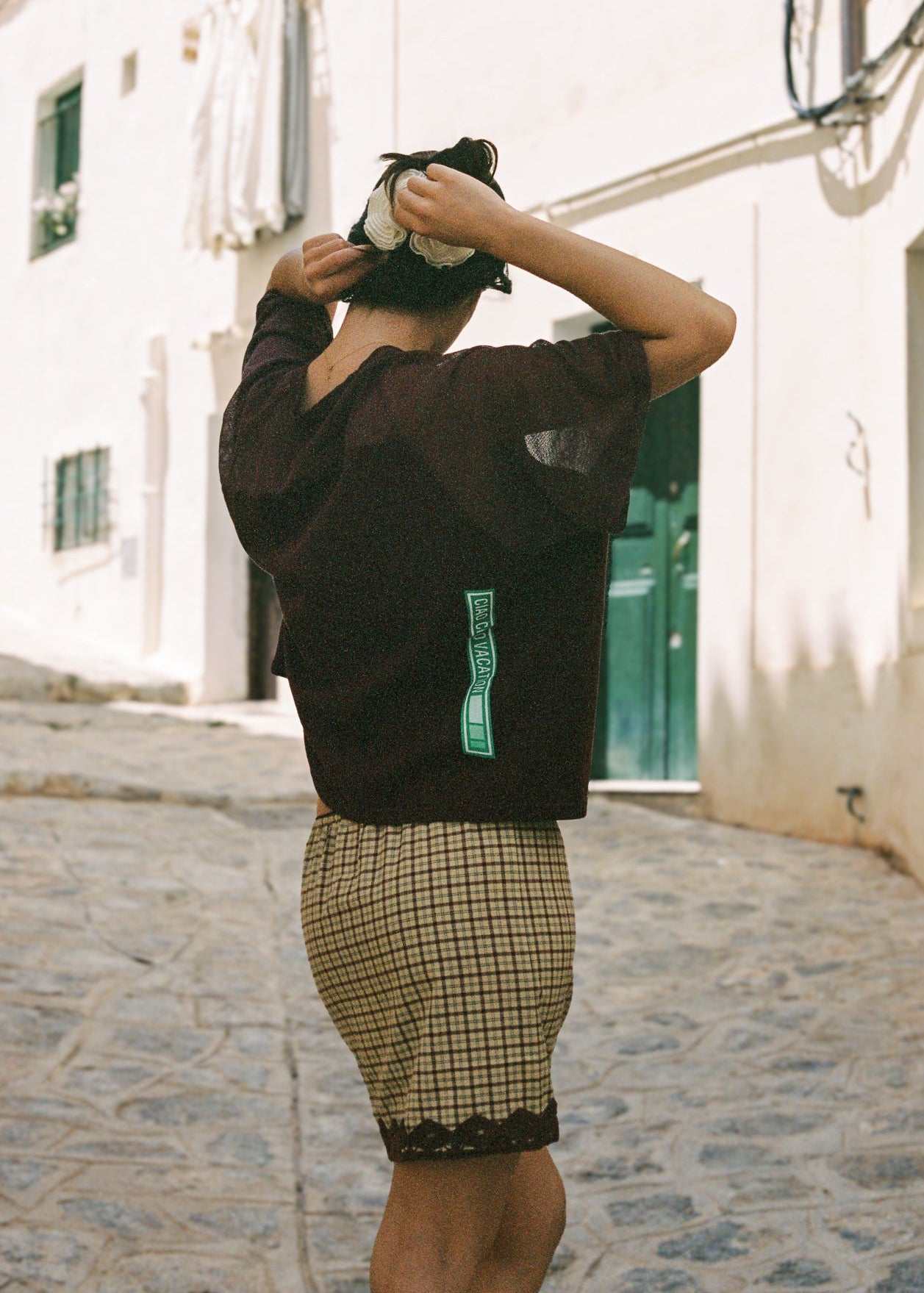 Person wearing a dark mesh tee with a green logo and plaid skirt walking on a stone street.