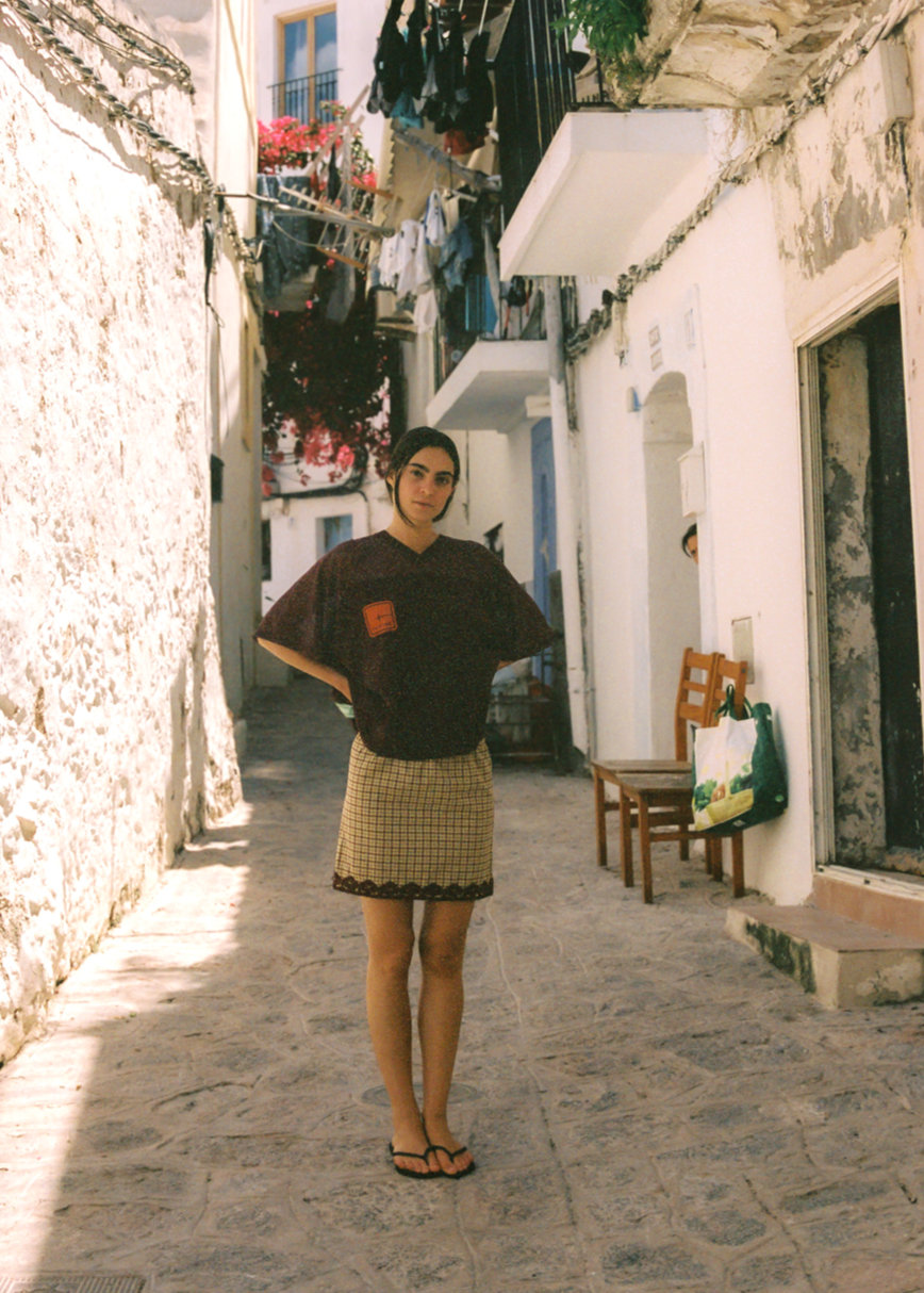 Woman standing in a narrow street wearing the tartan cropped mesh t-shirt and checkered skirt. 