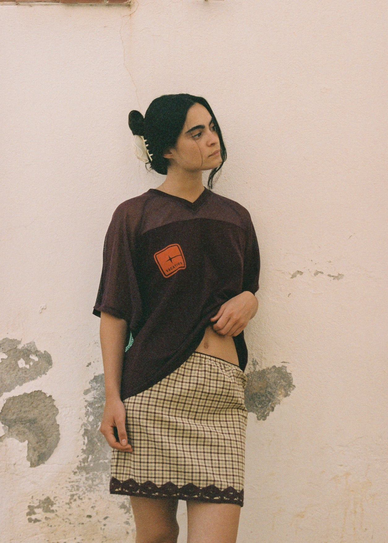 Person wearing a dark brown graphic oversized t-shirt with a red logo and a checkered tartan skirt against a textured wall.