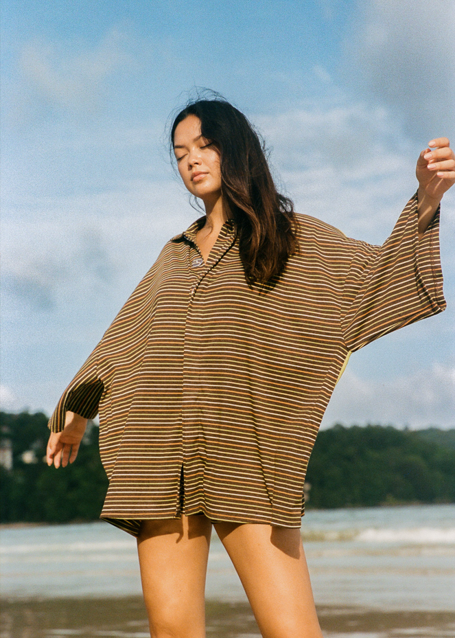 Woman standing on a beach with a brown oversized shirt with green, yellow and orange stripes 