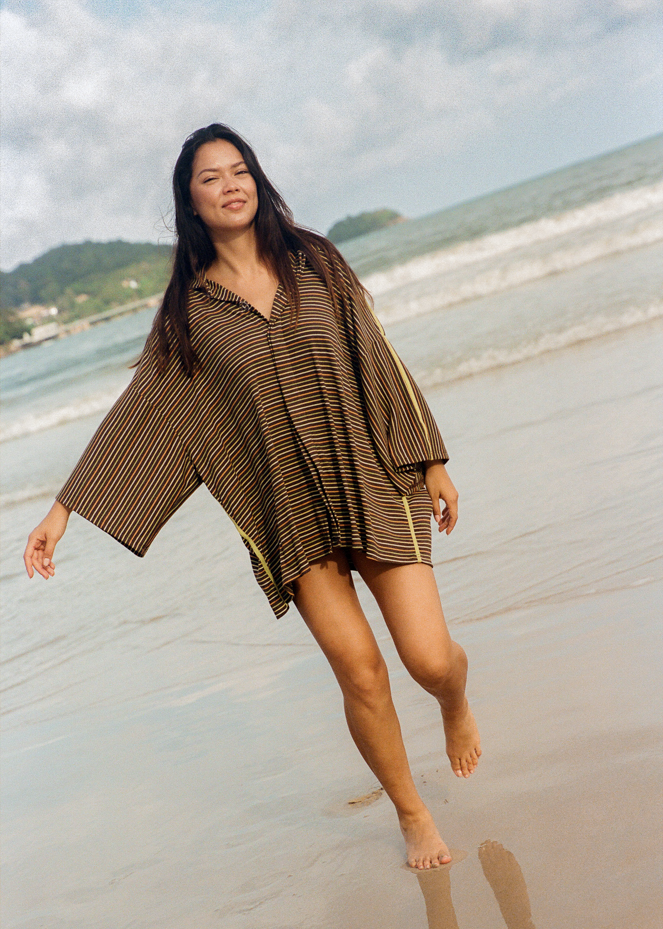 Woman standing on a beach with a brown oversized shirt with green, yellow and orange stripes 