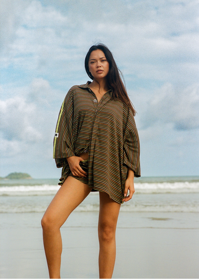 Woman standing on a beach with a brown oversized shirt with green, yellow and orange stripes 