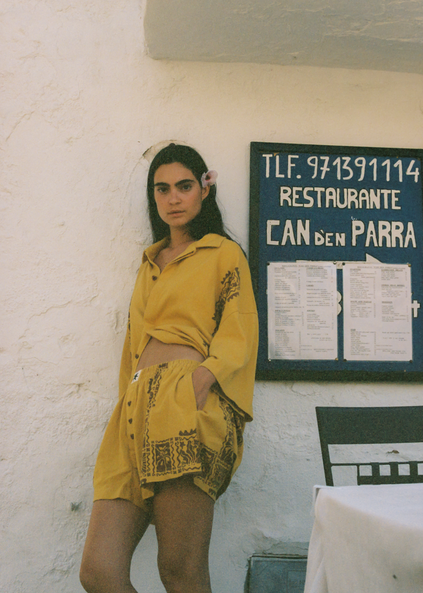 Woman in yellow and brown matching set outfit. Wearing a button up shirt and boxer shorts standing in front of a restaurant sign in Spanish.