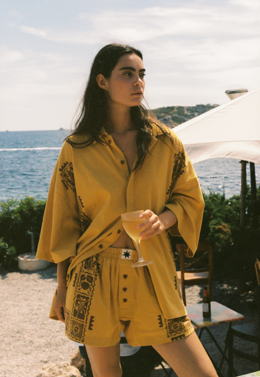 Woman in a yellow and brown matching set outfit by the sea. Wearing a oversized button up shirt and boxer shorts