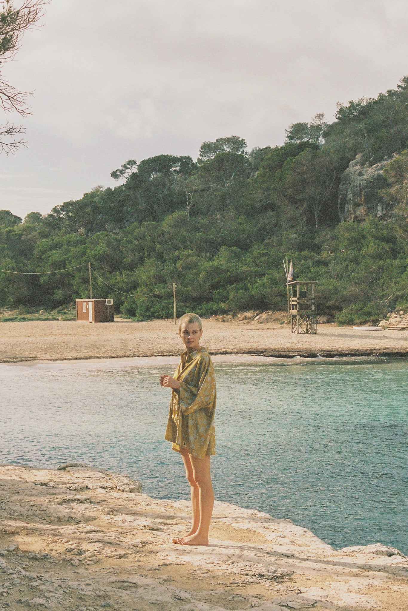 Person wearing a yellow satin outfit with botanical motifs standing by a body of water with buildings in the background.