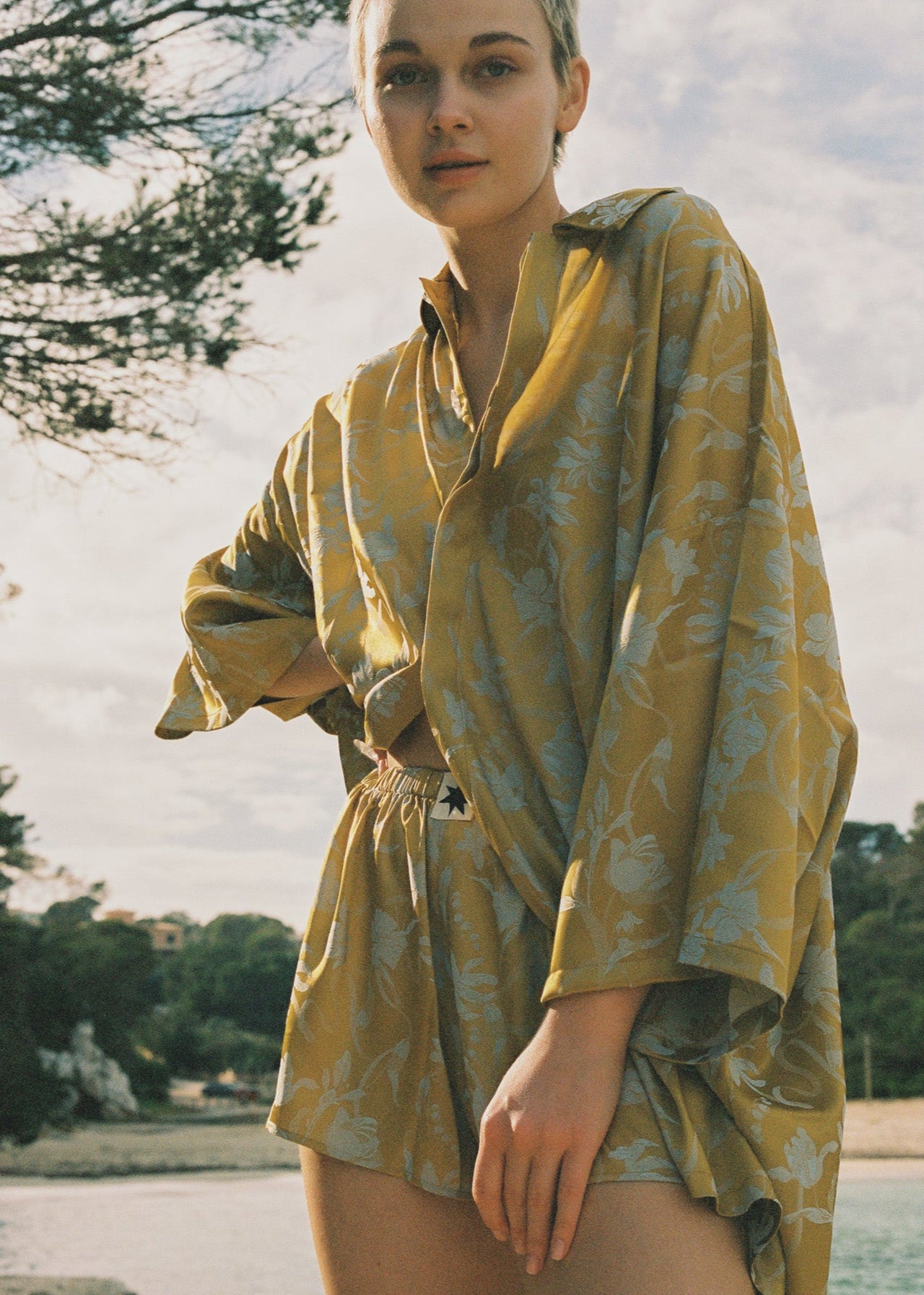 Person wearing a yellow satin outfit with botanical motifs standing by a body of water with buildings in the background.