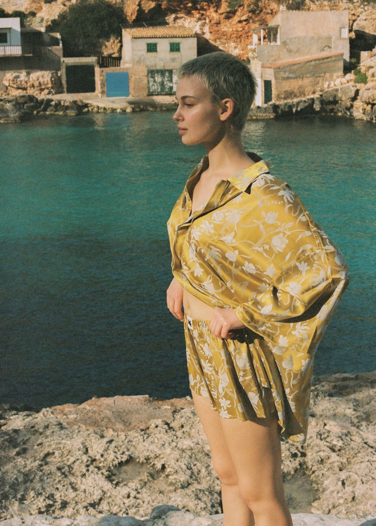 Person wearing a yellow satin outfit with botanical motifs standing by a body of water with buildings in the background.