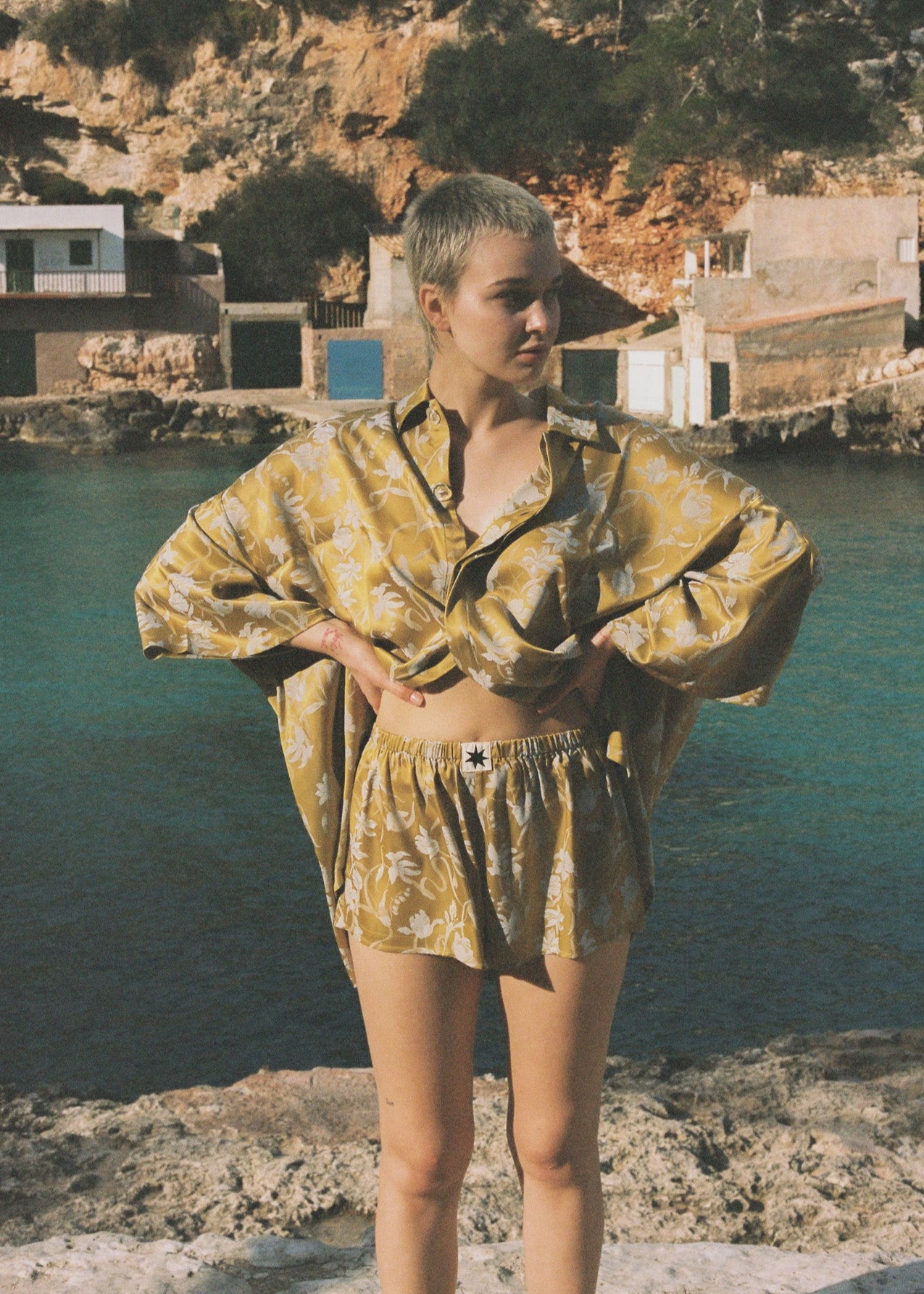 Person wearing a yellow satin outfit with botanical motifs standing by a body of water with buildings in the background.