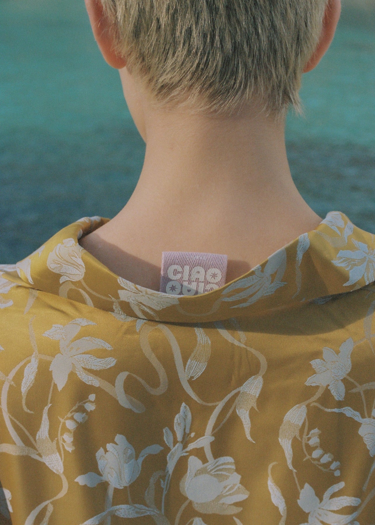 Person wearing a yellow floral satin shirt with a visible brand label, standing by a body of water.