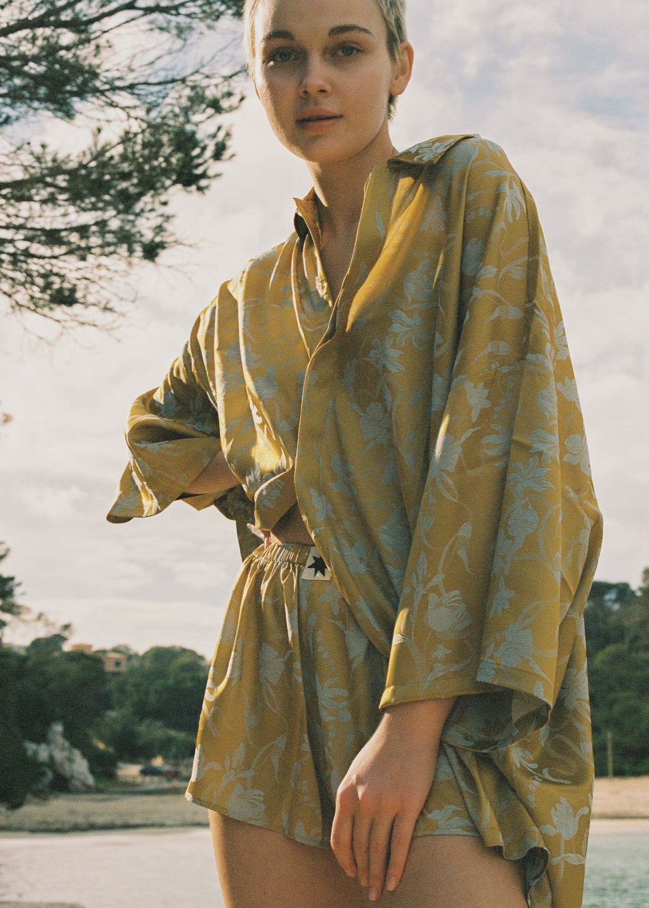 Woman wearing a yellow floral matching set outdoors with trees and sky in the background.