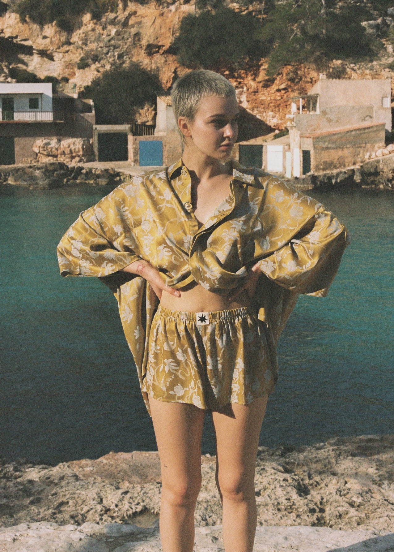 Person wearing a yellow floral patterned matching oversized set standing by a body of water with buildings in the background.