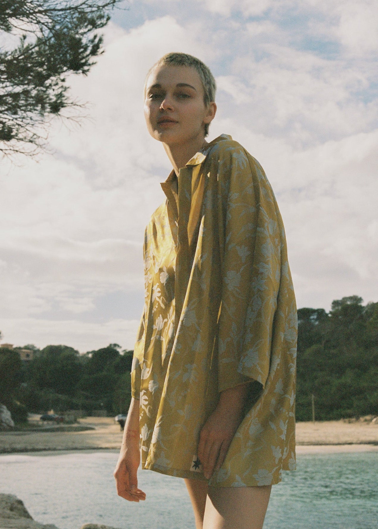 Person wearing a yellow floral patterned oversized button up shirt by a body of water with trees in the background