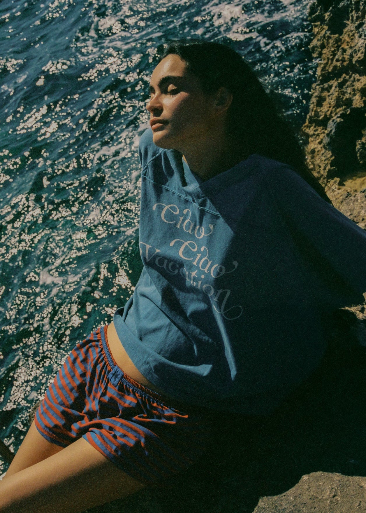 Person wearing a blue oversized t-shirt with text as well as a pair of striped shorts sitting by a body of water.