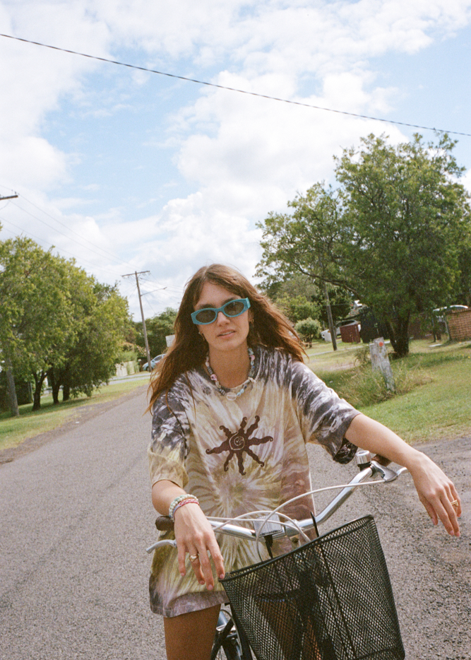 Woman riding a bicycle on a sunny day with a purple and green tie-dye box tee.